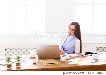 A woman sits at a desk in an office space. She looks focused as she writes notes while surrounded by crumpled papers. A laptop and notebooks are on the desk under bright light. A woman sits at a desk in an office space. She looks focused as she writes notes while surrounded by crumpled papers. A laptop and notebooks are on the desk under bright light. 143460002