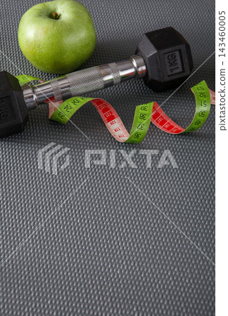A green apple sits next to a black dumbbell on a gray exercise mat. A measuring tape is curled around the dumbbell, showing numbers for fitness measurements. A green apple sits next to a black dumbbell on a gray exercise mat. A measuring tape is curled around the dumbbell, showing numbers for fitness measurements. 143460005