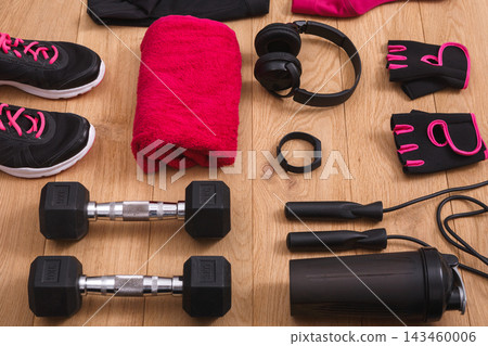 Different workout items are arranged on a wood surface. There are shoes, weights, gloves, a towel, a water bottle, and headphones ready for an exercise routine. Different workout items are arranged on a wood surface. There are shoes, weights, gloves, a towel, a water bottle, and headphones ready for an exercise routine. 143460006
