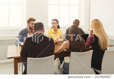 People gather around a table in a meeting room. They are engaged in conversation and sharing thoughts. It is daytime and sunlight enters through the windows. 143460007