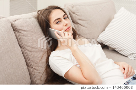 A young woman is lying on a couch while using her phone. She appears focused on the call, enjoying a moment of connection at home. Soft light fills the room. A young woman is lying on a couch while using her phone. She appears focused on the call, enjoying a moment of connection at home. Soft light fills the room. 143460008