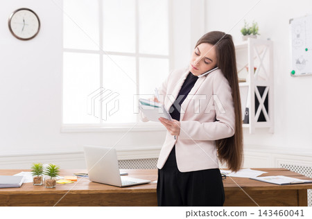 A business professional stands at a desk in an office while talking on the phone. She writes notes in a notepad with a laptop and documents present on the desk. A business professional stands at a desk in an office while talking on the phone. She writes notes in a notepad with a laptop and documents present on the desk. 143460041