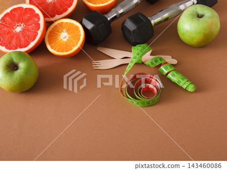 Fresh fruits including apples, oranges, and grapefruits are placed beside dumbbells. A measuring tape and wooden utensils are visible on a brown surface, related to fitness and health. Fresh fruits including apples, oranges, and grapefruits are placed beside dumbbells. A measuring tape and wooden utensils are visible on a brown surface, related to fitness and health. 143460086