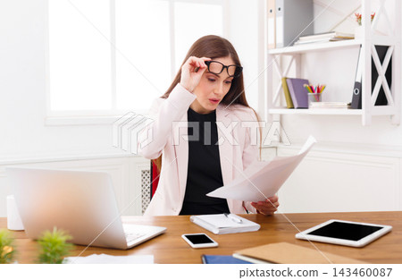A woman sits at a desk in a bright office. She holds paper in one hand and looks at it with focus. A laptop and two devices rest on the desk. Shelves in the background hold office supplies. A woman sits at a desk in a bright office. She holds paper in one hand and looks at it with focus. A laptop and two devices rest on the desk. Shelves in the background hold office supplies. 143460087