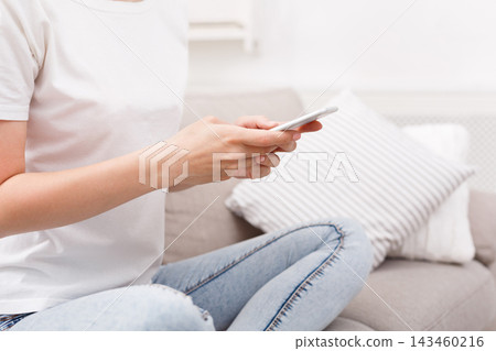 A person holds a smartphone while sitting on a sofa at home in a casual setting. The individual seems focused on the screen, enjoying the moment. A cozy pillow is beside them. A person holds a smartphone while sitting on a sofa at home in a casual setting. The individual seems focused on the screen, enjoying the moment. A cozy pillow is beside them. 143460216