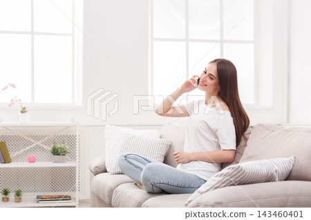 A woman is sitting on a comfortable sofa while talking on her phone. Natural light comes in from the windows, creating a warm atmosphere. She looks relaxed and engaged in conversation. A woman is sitting on a comfortable sofa while talking on her phone. Natural light comes in from the windows, creating a warm atmosphere. She looks relaxed and engaged in conversation. 143460401