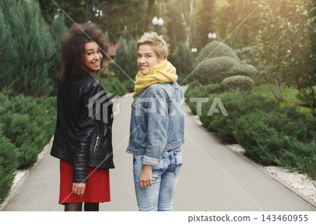 Two friends smile and look back while walking along a path in a garden. The area is filled with green plants and trees. They wear casual clothing and look happy in the setting. Two friends smile and look back while walking along a path in a garden. The area is filled with green plants and trees. They wear casual clothing and look happy in the setting. 143460955