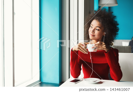 A woman sits in a cafe holding a cup of coffee while enjoying music through her earphones. She looks thoughtful as she gazes out the window. The cafe features a bright blue wall. 143461007