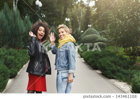 Two friends laugh and wave at each other while walking in a garden. They wear casual clothes and enjoy a sunny day surrounded by greenery. The scene captures their happiness. Two friends laugh and wave at each other while walking in a garden. They wear casual clothes and enjoy a sunny day surrounded by greenery. The scene captures their happiness. 143461108