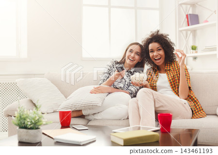 Two friends sit on a couch in a bright living room with snacks. One friend holds a remote and smiles while the other holds popcorn. They are relaxed and happy in their casual setting. Two friends sit on a couch in a bright living room with snacks. One friend holds a remote and smiles while the other holds popcorn. They are relaxed and happy in their casual setting. 143461193