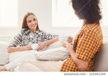 Two friends sit together on a sofa in a bright room. They smile and enjoy warm drinks while having a friendly conversation. The setting is simple and inviting, perfect for social time. Two friends sit together on a sofa in a bright room. They smile and enjoy warm drinks while having a friendly conversation. The setting is simple and inviting, perfect for social time. 143461194