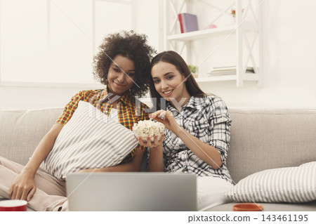 Two friends sit on a sofa in a cozy room. They are smiling while sharing a bowl of popcorn. A laptop is open in front of them as they watch a film together in the evening. Two friends sit on a sofa in a cozy room. They are smiling while sharing a bowl of popcorn. A laptop is open in front of them as they watch a film together in the evening. 143461195