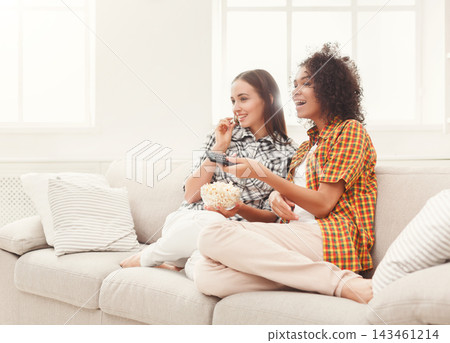 Two friends sit on a couch in a bright room. They are watching a movie and eating popcorn. One friend holds a remote. They smile and engage with the film. Two friends sit on a couch in a bright room. They are watching a movie and eating popcorn. One friend holds a remote. They smile and engage with the film. 143461214