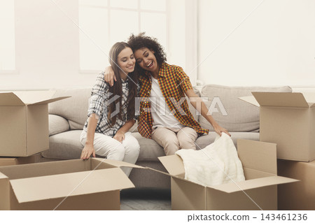 Two friends sit on a couch in a living room. They are smiling and relaxed while surrounded by cardboard boxes. It looks like they are moving into a new place. 143461236