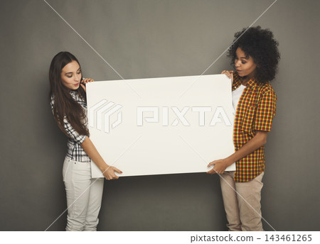 Two individuals stand side by side and hold a blank canvas. The scene takes place inside a studio with neutral walls. One person wears a checkered shirt and the other wears a plaid top. Two individuals stand side by side and hold a blank canvas. The scene takes place inside a studio with neutral walls. One person wears a checkered shirt and the other wears a plaid top. 143461265