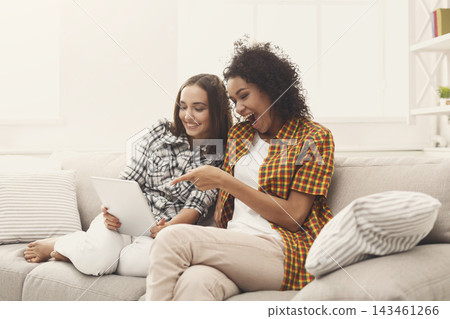 Two friends are sitting on a couch in a living room. They are looking at a tablet. One friend points at the screen while smiling. Sunlight is coming through the window. Two friends are sitting on a couch in a living room. They are looking at a tablet. One friend points at the screen while smiling. Sunlight is coming through the window. 143461266