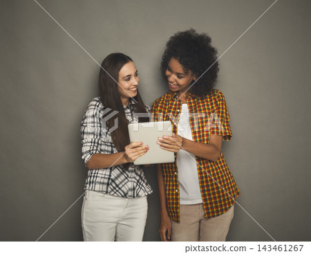 Two friends are looking at a tablet while smiling at each other. They are in a simple indoor space with a neutral background. The atmosphere is relaxed as they enjoy their time together. Two friends are looking at a tablet while smiling at each other. They are in a simple indoor space with a neutral background. The atmosphere is relaxed as they enjoy their time together. 143461267