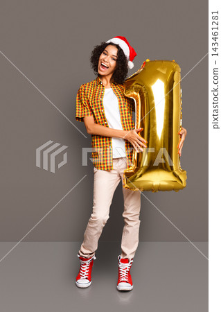 A child wears a festive outfit and a red hat while holding a large gold balloon in the shape of the number one. The background is simple. The child looks joyful and happy during the celebration. A child wears a festive outfit and a red hat while holding a large gold balloon in the shape of the number one. The background is simple. The child looks joyful and happy during the celebration. 143461281