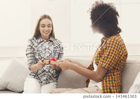 Two friends sit on a couch in a bright room. One friend hands a wrapped gift to the other. They are smiling and enjoying their time together. The atmosphere is friendly and relaxed. 143461303