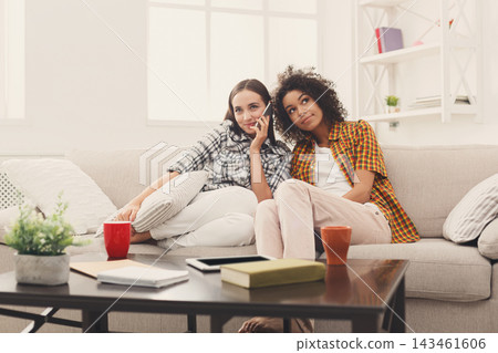 Two friends sit on a couch in a living room decorated with plants and books. They engage in conversation while holding drinks. Sunlight enters through the windows. Two friends sit on a couch in a living room decorated with plants and books. They engage in conversation while holding drinks. Sunlight enters through the windows. 143461606
