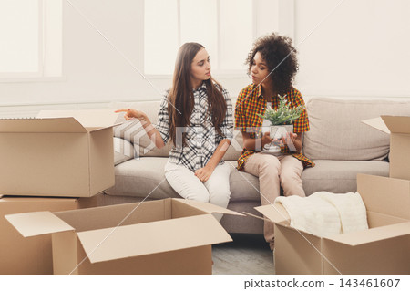 Two women sit on a couch in a room filled with cardboard boxes. They talk about their moving plans while one woman holds a small plant. The room has natural light coming in through windows. Two women sit on a couch in a room filled with cardboard boxes. They talk about their moving plans while one woman holds a small plant. The room has natural light coming in through windows. 143461607