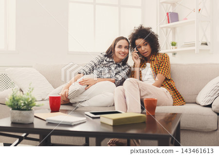 Two friends sit on a sofa in a cozy living room. They smile and chat while enjoying drinks. A table holds a book and devices. Bright light comes through the windows creating a warm atmosphere. Two friends sit on a sofa in a cozy living room. They smile and chat while enjoying drinks. A table holds a book and devices. Bright light comes through the windows creating a warm atmosphere. 143461613