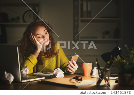 A woman sits at a desk with a laptop and a cup. She stares at her phone with a bored expression. Crumpled paper surrounds her in a dimly lit room filled with shelves. A woman sits at a desk with a laptop and a cup. She stares at her phone with a bored expression. Crumpled paper surrounds her in a dimly lit room filled with shelves. 143461878