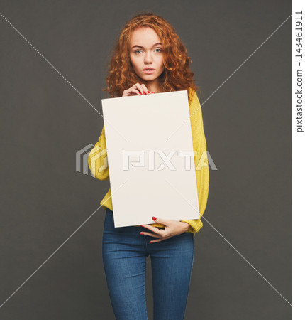 A person stands in a studio holding a blank canvas. The setting has a simple background. The individual looks directly at the camera while dressed in casual clothing. A person stands in a studio holding a blank canvas. The setting has a simple background. The individual looks directly at the camera while dressed in casual clothing. 143461911