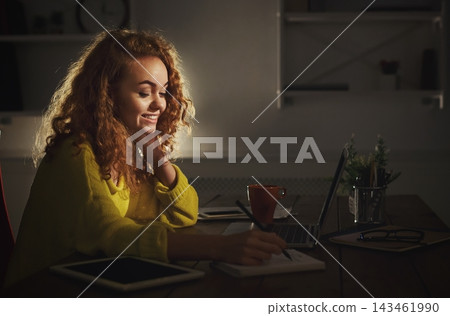 A young woman sits at a desk in a well-lit room. She is smiling and writing in a notebook while using a laptop. Surrounding her are plants and devices. A young woman sits at a desk in a well-lit room. She is smiling and writing in a notebook while using a laptop. Surrounding her are plants and devices. 143461990