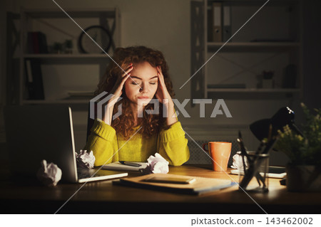 A young woman sits at a desk in a dimly lit room. She is focused and stressed as she works on her collage. There are crumpled papers around her, and a laptop is open next to her. A young woman sits at a desk in a dimly lit room. She is focused and stressed as she works on her collage. There are crumpled papers around her, and a laptop is open next to her. 143462002