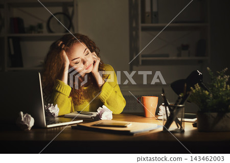 A woman sits at a desk late at night, looking thoughtful while surrounded by a laptop, coffee cup, and crumpled paper. The room has bookshelves and a lamp providing warm light. A woman sits at a desk late at night, looking thoughtful while surrounded by a laptop, coffee cup, and crumpled paper. The room has bookshelves and a lamp providing warm light. 143462003