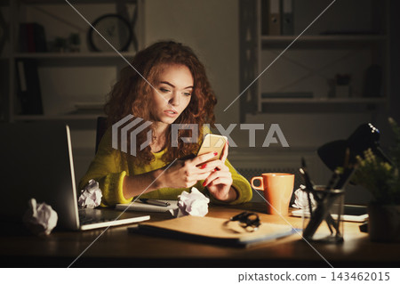 A woman sits at a desk in a dark room while looking at her smartphone. Papers are crumpled on the desk. A laptop and a mug are nearby. A lamp provides light for her work. A woman sits at a desk in a dark room while looking at her smartphone. Papers are crumpled on the desk. A laptop and a mug are nearby. A lamp provides light for her work. 143462015
