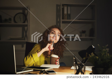A woman sits at a table with a laptop, coffee cup, and notebook. She looks thoughtful while holding a pen. The room has soft lighting and shelves in the background. A woman sits at a table with a laptop, coffee cup, and notebook. She looks thoughtful while holding a pen. The room has soft lighting and shelves in the background. 143462016