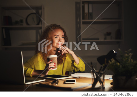 A woman sits at a desk holding a cup of coffee. She smiles while working on her laptop. The room is dark with a lamp providing light. Books and a plant are nearby. 143462063
