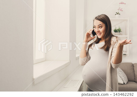 A pregnant woman stands near a window in a living room. She smiles while talking on her phone. Her hand gestures show excitement. A couch and plants are in the background. 143462292