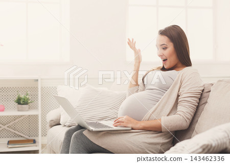 A pregnant woman sits on a light couch with a laptop in front of her. She looks surprised and happy as she raises her hand to express her excitement. Sunlight comes through the windows. A pregnant woman sits on a light couch with a laptop in front of her. She looks surprised and happy as she raises her hand to express her excitement. Sunlight comes through the windows. 143462336