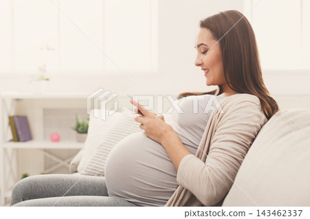 A woman sits comfortably on a couch, engaged with her smartphone. She is in a light room with decorative items around her. This scene captures a moment during her pregnancy at home. 143462337