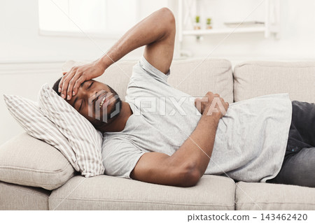 A man lies on a couch in a living room, holding his head with one hand and his stomach with the other. The setting has bright light coming in through a window. A man lies on a couch in a living room, holding his head with one hand and his stomach with the other. The setting has bright light coming in through a window. 143462420