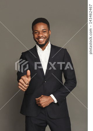 A man stands in front of a grey background wearing a suit. He smiles and gives a thumbs up. This is part of a collection of professional portraits showcasing different expressions and attitudes. 143462467