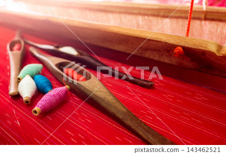 Traditional wooden weaving shuttle and thread on red handmade fabric, textile loom tools used for cloth production. Artisan craftsmanship, cultural heritage and weaving tradition. Fabric making. Traditional wooden weaving shuttle and thread on red handmade fabric, textile loom tools used for cloth production. Artisan craftsmanship, cultural heritage and weaving tradition. Fabric making. 143462521