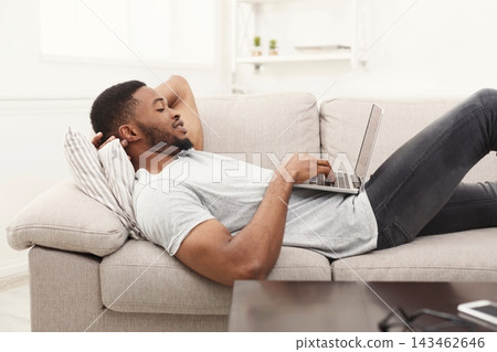 A young man is lying back on a light sofa with a laptop in his hands. He appears focused on the screen. The living room has simple decor and soft light coming through windows. A young man is lying back on a light sofa with a laptop in his hands. He appears focused on the screen. The living room has simple decor and soft light coming through windows. 143462646