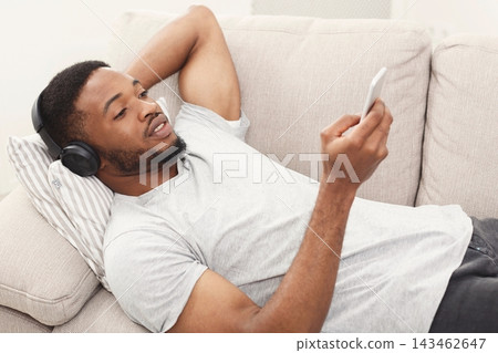 A person rests on a couch with headphones on, looking at a phone. The setting is indoors with soft furnishings. This scene captures a casual moment of leisure and enjoyment. 143462647