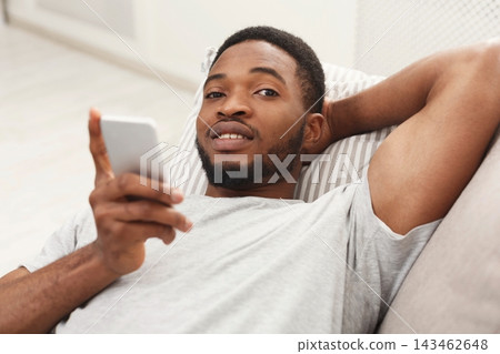 A man is lying on a couch in a living room. He is relaxing and looking at his smartphone. The room is bright and has a simple design. He is smiling while browsing. A man is lying on a couch in a living room. He is relaxing and looking at his smartphone. The room is bright and has a simple design. He is smiling while browsing. 143462648