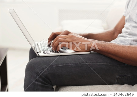 A person sits on a couch with a laptop. Their hands are on the keyboard as they type. The room has natural light coming in through the window, creating a warm atmosphere. 143462649