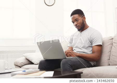 A man is sitting on a couch in a light-filled room. He focuses on his laptop while typing. The setting shows a clock on the wall, giving a sense of home office work during the day. A man is sitting on a couch in a light-filled room. He focuses on his laptop while typing. The setting shows a clock on the wall, giving a sense of home office work during the day. 143462650