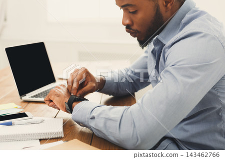 A man sits at a wooden desk looking at his smartwatch. A laptop is open beside him, and there are notepads, pens, and sticky notes on the table. This takes place in a bright office. 143462766