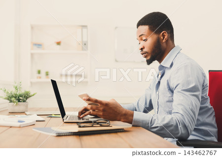 A man sits at a wooden desk in a modern office space. He uses a laptop while holding a phone. The desk has some plants and office supplies. Natural light fills the room. A man sits at a wooden desk in a modern office space. He uses a laptop while holding a phone. The desk has some plants and office supplies. Natural light fills the room. 143462767