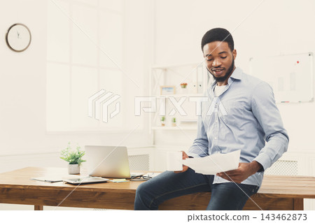 A man is sitting at a desk in a bright office. He holds papers in one hand and looks at them. There is a laptop and a small plant on the table. The space is well lit. A man is sitting at a desk in a bright office. He holds papers in one hand and looks at them. There is a laptop and a small plant on the table. The space is well lit. 143462873