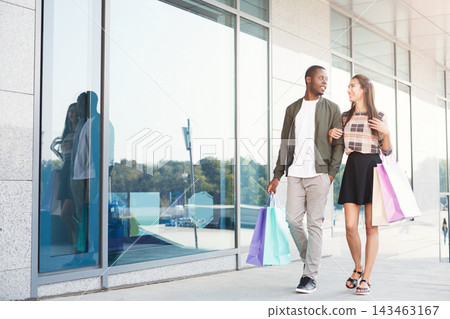 A couple is walking side by side outside a retail building. They hold shopping bags in their hands. The building has large windows that reflect their image. It is daytime and they appear happy. A couple is walking side by side outside a retail building. They hold shopping bags in their hands. The building has large windows that reflect their image. It is daytime and they appear happy. 143463167