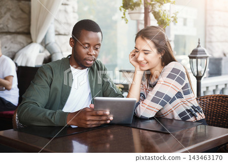 Two individuals are seated at a table in a cafe, focused on a tablet. They share smiles and appear engaged in their activity. Sunlight filters through the outdoor setting. Two individuals are seated at a table in a cafe, focused on a tablet. They share smiles and appear engaged in their activity. Sunlight filters through the outdoor setting. 143463170